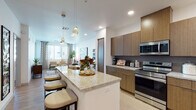 This well-appointed single-floor residence showcases a contemporary design with a cohesive blend of warm wood tones, granite surfaces, and light hardwood flooring throughout. The open-concept kitchen is a standout feature, equipped with full-height flat-front cabinetry in a rich wood finish, a textured tile backsplash, stainless steel appliances including a range, microwave, refrigerator, and dishwasher, and an extended granite-topped island with bar seating and pendant lighting overhead. The kitchen flows seamlessly into the dining and living areas, creating an expansive, connected layout ideal for entertaining. The living room is bright and welcoming, with multiple windows dressed in patterned drapes that invite natural light while offering privacy, complemented by a ceiling fan overhead. Two bedrooms provide comfortable retreats, each with plush carpeting, ceiling fans, and windows with window treatments. The primary bathroom features a lengthy dual-sink granite vanity with wood cabinetry, a large mirror with overhead lighting, and direct access to the primary bedroom. Clean white walls, recessed lighting, and consistent material choices throughout contribute to a polished, modern ambiance.