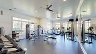 Located at Madison Park in Huntsville, Alabama, this fitness center offers a bright and well-appointed space for residents to pursue their wellness goals. The room features a clean, open layout with durable rubber flooring designed to support a full range of exercise activities. Large, multi-pane windows line the walls on multiple sides, allowing abundant natural light to fill the space while providing pleasant outdoor views of the surrounding greenery. Ceiling fans with brushed-metal finishes provide air circulation overhead, complementing the recessed lighting and crown molding details that lend a polished, residential feel to the space. The cardio zone is well-stocked with treadmills, elliptical trainers, and stationary bikes, while a dedicated strength area includes cable machines, a multi-function weight station, adjustable benches, and a comprehensive dumbbell rack with medicine balls. A wall-length mirror expands the sense of space and supports proper form during workouts. A bold, geometric accent mural in yellow, black, and white adds a dynamic visual focal point to the free weights area. Wall-mounted televisions provide entertainment options throughout. French doors with glass panes contribute to the airy, open character of this thoughtfully designed community fitness facility.