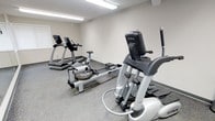This single-floor fitness room offers a focused workout environment outfitted with a selection of cardio equipment to accommodate a range of exercise preferences. The space features True-brand elliptical and treadmill machines, along with a WaterRower rowing machine, providing users with diverse low-impact and aerobic training options. The room is finished with neutral beige walls and durable speckled flooring, which is both practical and easy to maintain. Overhead fluorescent lighting ensures consistent, bright illumination throughout the space, making it suitable for use at any time of day. The layout is thoughtfully arranged to allow users comfortable access to each piece of equipment without crowding. The clean, minimalist design keeps the focus on functionality, with no unnecessary décor to distract from the purpose of the room. The space is well-suited for individual workouts and provides a convenient fitness option without the need to travel to an off-site gym facility.