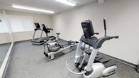 This single-floor fitness room offers a focused workout environment outfitted with a selection of cardio equipment to accommodate a range of exercise preferences. The space features True-brand elliptical and treadmill machines, along with a WaterRower rowing machine, providing users with diverse low-impact and aerobic training options. The room is finished with neutral beige walls and durable speckled flooring, which is both practical and easy to maintain. Overhead fluorescent lighting ensures consistent, bright illumination throughout the space, making it suitable for use at any time of day. The layout is thoughtfully arranged to allow users comfortable access to each piece of equipment without crowding. The clean, minimalist design keeps the focus on functionality, with no unnecessary décor to distract from the purpose of the room. The space is well-suited for individual workouts and provides a convenient fitness option without the need to travel to an off-site gym facility.