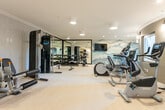 This fitness center is thoughtfully designed to accommodate a full range of workout activities across its single-level layout. The space is divided into distinct functional zones, each equipped with professional-grade machinery. The cardio area features multiple treadmills and elliptical machines, all positioned facing a dramatic watercolor-style mountain landscape mural that spans an entire accent wall. A wall-mounted flat-screen television is centered above the mural, providing entertainment during workouts.

The strength training zone is anchored by a large, black-framed floor-to-ceiling mirror that visually expands the space while allowing users to monitor their form. A full dumbbell rack and adjustable weight bench are positioned nearby, complemented by a cable machine and leg press unit for comprehensive resistance training.

The flooring throughout is light-toned and cushioned, offering comfort underfoot. Textured light grey wallcovering lines several walls, contributing to the refined, neutral aesthetic. Flush-mount and recessed ceiling lights provide consistent, even illumination across the entire space.

Additional features include built-in shelving for storage, a small refreshment counter with a beverage refrigerator, and interior windows that offer views into an adjacent stairwell, enhancing the sense of openness within this well-equipped fitness facility.