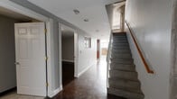 Located at 1429 Park St in Hartford, CT, this two-floor urban loft showcases a compelling mix of industrial heritage and contemporary design. The lower level features a wide corridor with dark hardwood floors, recessed lighting, and classically trimmed doorways that open to versatile rooms with clean grey walls and stained concrete flooring. Large multi-pane windows flood the entry and common areas with natural light, while an exposed brick accent wall adds raw, historic character. A staircase with a warm wood handrail connects the two floors, maintaining an open, flowing feel throughout. The well-appointed kitchen features rich wood cabinetry, dark granite countertops, a checkered tile floor, and a full suite of stainless steel appliances including a range, microwave, refrigerator, and dishwasher. The upper level reveals soaring exposed concrete ceilings with visible ductwork and structural columns — hallmark loft details that lend an authentic industrial aesthetic. The bathroom is distinctively designed, featuring a tiled soaking tub, warm hardwood floors, and a striking wall-mounted glass sink vanity with chrome hardware. A spacious walk-in closet with wire shelving provides ample storage. Throughout both floors, the interplay of concrete, brick, hardwood, and modern finishes creates a bold, architecturally rich environment.