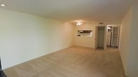 This single-floor residential space offers a straightforward and functional layout designed for comfortable everyday living. The main living area is generously sized, finished with wall-to-wall carpet flooring in a neutral beige tone that lends warmth to the room. A ceiling fan centered overhead provides both lighting and ventilation, contributing to the room's practicality. The walls are painted in a light, neutral tone, creating a clean and open atmosphere throughout.

A standout feature of the living area is the pass-through window that connects directly to the kitchen, allowing for easy interaction between the two spaces. The kitchen, visible through the pass-through, features dark wood cabinetry and a light-colored countertop, offering a classic contrast. A textured ceiling adds a subtle architectural detail to the space.

Adjacent to the living area is a dedicated closet with a rod and shelf, providing practical storage solutions. The overall ambiance of the space is calm and neutral, making it a versatile canvas for a variety of interior styles. The single-floor layout ensures ease of movement and accessibility throughout, while the open feel of the main living area makes the space feel welcoming and adaptable.