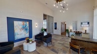 This contemporary single-floor space serves as a polished community lobby or leasing center, thoughtfully designed to make a strong first impression. The interior is anchored by dramatically high ceilings that amplify the sense of openness throughout the main lounge area. Large floor-to-ceiling windows draped with sheer white curtains flood the space with natural light while offering views of the surrounding landscaping and community exterior.

The lounge features a refined seating arrangement of charcoal upholstered armchairs gathered around a modern round white coffee table. A textured accent wall with a deep cobalt blue recessed panel showcases a vibrant abstract painting, flanked by sleek wall sconces that add a warm, ambient glow. The flooring consists of large-format mixed-tone stone tiles that lend an earthy yet sophisticated character to the space.

A glass-paneled partition wall separates the lounge from a private office area, where a substantial mid-century modern wood desk is positioned to take full advantage of the surrounding natural light. Additional areas include a hallway leading to secondary rooms and a credenza with mirrored detailing that adds elegance near an informational display board. A striking sputnik-style brass chandelier crowns the main lounge, completing the stylish, contemporary aesthetic throughout.