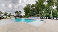 Located at 1215 Evolve Way in Greenville, North Carolina, this outdoor amenity space is designed as a resort-style pool area that offers a comprehensive recreational experience. The centerpiece is a generously sized, multi-section swimming pool with crystal-clear blue water, entry handrails, and clearly marked depth indicators, accommodating both leisure and active swimmers. The expansive pool deck is finished in textured concrete and paver surfaces, providing ample space for relaxation and socializing. Rows of modern chaise lounge chairs with blue cushions are arranged throughout the deck, offering comfortable seating for sunbathers. A distinctive pergola structure provides shaded lounging areas along one side of the pool, while a charming gazebo sits at the far end of the complex, adding architectural character to the space. A covered pavilion building with board-and-batten siding and a metal roof anchors the facility, featuring large windows and a covered porch that overlooks the pool area. The entire space is enclosed by a sleek black iron fence with a gated entry, ensuring safety and security. Mature trees form a lush green backdrop, creating a sense of natural seclusion and a serene outdoor ambiance throughout the complex.