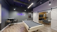 Located at 2901 Connecticut Avenue Northwest in Washington, DC, this amenity lounge is a thoughtfully designed communal space that blends industrial aesthetics with modern comfort. The open-plan layout is unified by light wood-look flooring and an exposed black ceiling with visible ductwork and pendant lighting, lending the space an urban, loft-inspired character.

At the heart of the main area sits a full-size pool table, serving as the focal point for social gatherings. A neon DC skyline sign adds a local, artistic touch to the dark-painted accent walls. Sliding barn doors — both classic white-paneled and frosted glass varieties — connect distinct zones throughout the space, offering flexible separation between areas.

A dedicated lounge zone features gray upholstered sofas centered around a flat-screen television, creating a relaxed viewing area. Nearby, a bar-height counter with sleek stools provides a casual spot for conversation or independent work, while a collection of framed black-and-white photography adds a gallery-like quality to the walls. A small bistro table with chairs offers additional seating for intimate gatherings.

Structural columns and open sightlines throughout contribute to the spacious feel of this well-appointed amenity room, making it a versatile hub for recreation and community engagement.