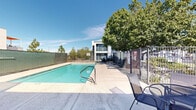 This outdoor pool area offers a well-appointed recreational space designed for relaxation and leisure. At the center of the space is a generously sized swimming pool with clear, blue-green water, accessible via stainless steel handrails and step entries. The pool is surrounded by a broad, light-colored concrete deck that provides ample room for movement and sunbathing.

A variety of seating options are arranged around the perimeter, including lounge chairs and patio furniture, offering guests comfortable spaces to unwind poolside. Mature shade trees are positioned throughout the area, contributing natural coverage and a pleasant outdoor ambiance. The layout takes full advantage of abundant natural light and open skies, creating a bright and inviting atmosphere.

The pool area is fully enclosed by a combination of iron fencing and privacy screening, ensuring both security and seclusion. An adjacent low-rise building with direct access doors is visible along one side, likely housing restrooms or utility facilities to support the pool area. The surrounding structures reflect a clean, modern architectural style with neutral exterior finishes and large windows.

Overall, this outdoor pool space is practical, attractive, and well-maintained, making it a functional amenity suited for a variety of users seeking a refreshing outdoor retreat.