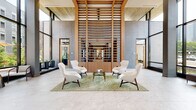 Located at 7970 Maitland St in McLean, Virginia, this striking commercial lobby showcases a sophisticated blend of natural materials and contemporary design. The grand entrance features floor-to-ceiling glass curtain walls and a prominent revolving glass door, flooding the expansive space with abundant natural light and offering unobstructed views of the surrounding urban landscape.

At the heart of the lobby stands a distinctive live-edge wood reception desk set against a dramatic bookmatched marble accent wall, creating a bold focal point upon entry. Overhead, warm wood-slat ceiling panels add texture and depth, complementing the polished large-format stone tile flooring that extends throughout.

A central lounge area features a curated seating arrangement of mid-century modern chairs arranged around a glass coffee table atop a sculptural green area rug, set beneath a soaring double-height wood-slat architectural screen that serves as a dramatic interior partition. Floor-to-ceiling glazing on the rear façade frames views of a landscaped outdoor courtyard furnished with seating and market umbrellas.

A corridor lined with steel-framed glass partitions leads to private office spaces and conference rooms, with curated artwork displayed along warm wood-paneled walls. The overall ambiance is one of refined elegance, balancing natural warmth with sleek, modern professionalism.