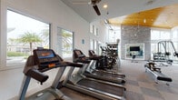 Located at 1040 Jamison Loop in Kissimmee, Florida, this single-story fitness center is designed to offer a comprehensive and motivating workout environment. The main exercise area is defined by dramatically high ceilings with a striking wood-paneled vaulted section and an exposed stone accent column, lending the space a modern yet warm architectural character. Large floor-to-ceiling windows and clerestory windows flood the interior with natural light while offering views of the surrounding palm-lined grounds.

The cardio zone features a row of Matrix treadmills and elliptical trainers positioned along the windowed walls, complemented by ceiling fans that enhance airflow throughout. The strength training area is outfitted with a multi-station cable machine, weight benches, free weights, and a dumbbell rack, all arranged on durable checkered carpet tile flooring. Full-length mirrors line multiple walls, maximizing both the sense of space and workout functionality.

A dedicated cycling studio is set apart from the main floor, featuring spin bikes, wood-look flooring, a coffered ceiling with recessed lighting, and floor-to-ceiling mirrors — creating an immersive space ideal for group or individual cycling sessions. The facility is well-lit, thoughtfully arranged, and designed to accommodate a wide range of fitness activities in a clean, modern setting.