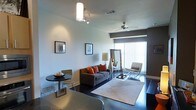 This single-floor space showcases a thoughtfully designed open-concept living area with a clean, modern aesthetic. Dark hardwood floors anchor the room, providing a striking contrast against the light-toned walls and creating a sleek, contemporary feel throughout.

The living area is comfortably appointed with a gray sectional sofa accented by vibrant orange throw pillows, adding warmth and a pop of color to the neutral palette. A large floor mirror leans against one wall, enhancing the sense of depth and amplifying natural light. A stylish floor lamp and a cushioned accent chair complete the seating arrangement.

Large sliding glass doors flood the space with natural light and offer access to what appears to be an outdoor area, contributing to an airy, open ambiance. Vertical blinds provide privacy and light control as needed.

A ceiling fan with a light fixture ensures year-round comfort, while a combination pendant and track lighting fixture near the dining area adds focused illumination. A compact round dining table with metal chairs defines a casual dining nook. A light-toned area rug delineates the living space, softening the room and tying the furnishings together cohesively.