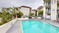 Located at 3216 Locust Ave in Long Beach, California, this outdoor amenity space offers a well-appointed communal pool area that embodies classic Southern California living. The centerpiece is a generously sized swimming pool with clear, blue-green water, framed by a smooth, light-colored concrete deck that provides ample space for lounging and entertaining. Sun loungers are positioned poolside, offering comfortable spots to relax and take in the open-air surroundings. A dedicated outdoor BBQ station with a large gas grill and adjacent seating area makes the space ideal for gatherings and al fresco dining. The area is framed by mature palm trees and lush greenery, enhancing the resort-like atmosphere. A perimeter wall with landscaped plantings provides privacy while still allowing views of the surrounding neighborhood skyline. The adjacent multi-story building features a Spanish-influenced architectural style, characterized by terracotta-tiled rooflines, stucco exteriors, and private balconies that overlook the courtyard. A gated entry adds a layer of security to the shared space. Together, these elements create a vibrant and functional outdoor retreat well-suited to the Long Beach climate.