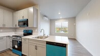 This thoughtfully designed two-bedroom, one-bathroom residence offers a clean, modern aesthetic across a single floor. The open kitchen is a standout feature, equipped with shaker-style cabinetry in a soft gray finish, white countertops, stainless steel appliances including a range, over-the-range microwave, and refrigerator, plus a dishwasher. Wood-look luxury vinyl plank flooring runs throughout the main living and kitchen areas, while a peninsula provides added workspace and a natural transition into the living room. A window in the living area frames scenic views of rolling hills and colorful autumn foliage, filling the space with natural light. A wall-mounted mini-split unit provides efficient climate control. Both bedrooms feature plush carpeting and large windows that capture the same picturesque outdoor views. The primary bedroom offers a walk-in closet with wire shelving for organized storage. The full bathroom is finished with wood-look flooring, a single-sink vanity with shaker cabinetry, a large mirror, and a walk-in shower with grab bars. An in-unit laundry area and two closets enhance everyday functionality. The neutral color palette and modern finishes throughout create a cohesive, inviting living environment.