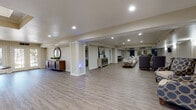 This thoughtfully designed amenity floor offers a comprehensive range of communal spaces, all unified by light-toned luxury vinyl plank flooring and a warm, neutral color palette. The expansive open lounge area serves as the heart of the floor, featuring decorative white columns, recessed lighting, and stylish furnishings including wingback chairs, a sofa arrangement, and accent pieces such as a round mirror above a wooden sideboard. A skylit entry foyer welcomes visitors with French doors that invite natural light, a statement console table, and an elegant coffered ceiling detail. The dining area accommodates multiple seating groups with granite-topped tables and coordinating teal and gray upholstered chairs, enhanced by soft wall sconces and recessed lighting. A dedicated media room provides tiered rows of plush two-toned seating facing a wall-mounted flat-screen television, ideal for group viewings. The fitness room is equipped with a treadmill and elliptical trainer, with floor-to-ceiling mirrors amplifying the space. Additional seating alcoves with patterned lounge chairs offer quieter retreats. Throughout, crown molding, clean white millwork, and consistent finishes create a polished, cohesive environment suited for both relaxation and social engagement.