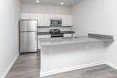 This single-floor unit in Warren, Michigan offers a clean, modern aesthetic defined by consistent finishes and an open layout. Light-toned wood-look flooring runs throughout the space, lending warmth and visual continuity from room to room.

The kitchen serves as a central highlight, featuring white shaker-style cabinetry, speckled granite countertops, and a full suite of stainless steel appliances, including a refrigerator, electric range, and over-the-range microwave. A peninsula island with a built-in sink and matching granite surface provides additional workspace and potential seating. Recessed lighting illuminates the kitchen efficiently.

The living area is open and versatile, benefiting from natural light through a window with blinds. A sliding barn door adds a contemporary architectural detail and connects adjoining spaces.

The bedroom includes a generously sized closet with wire shelving, offering practical storage. The bathroom is well-appointed with a granite-topped white vanity, a full tub-shower combination with built-in shelving, and a large mirror. The neutral color palette is maintained throughout, creating a cohesive and calm atmosphere.

Overall, the unit presents a well-designed, move-in-ready space with modern finishes and functional layout suited for comfortable everyday living.