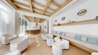 This single-floor space showcases a refined coastal aesthetic defined by a striking coffered ceiling system of light natural wood beams set against crisp white panels, creating visual depth and warmth throughout. The open layout is anchored by a central oval bar with a white stone countertop and a ribbed wood base, surrounded by mid-century-style wooden counter stools. Overhead, a curated mix of woven rattan and geometric pendant lights reinforces the space's relaxed yet sophisticated character.

The lounge area features a long built-in banquette upholstered in soft blush tones, accented with aqua and patterned throw pillows, and backed by a white shiplap wall with a floating natural wood display shelf. Sculptural white lounge chairs and organic-form coffee tables are arranged throughout, encouraging social interaction. Large floor-to-ceiling windows and glass doors flood the interior with natural light and frame views of palm trees and a pool terrace beyond. A decorative chain-link partition screen adds an artistic layer of transparency between the bar zone and the outdoors. Light wide-plank wood flooring and neutral tile ground the space in an airy, resort-like ambiance.
