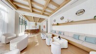 This single-floor space showcases a refined coastal aesthetic defined by a striking coffered ceiling system of light natural wood beams set against crisp white panels, creating visual depth and warmth throughout. The open layout is anchored by a central oval bar with a white stone countertop and a ribbed wood base, surrounded by mid-century-style wooden counter stools. Overhead, a curated mix of woven rattan and geometric pendant lights reinforces the space's relaxed yet sophisticated character.

The lounge area features a long built-in banquette upholstered in soft blush tones, accented with aqua and patterned throw pillows, and backed by a white shiplap wall with a floating natural wood display shelf. Sculptural white lounge chairs and organic-form coffee tables are arranged throughout, encouraging social interaction. Large floor-to-ceiling windows and glass doors flood the interior with natural light and frame views of palm trees and a pool terrace beyond. A decorative chain-link partition screen adds an artistic layer of transparency between the bar zone and the outdoors. Light wide-plank wood flooring and neutral tile ground the space in an airy, resort-like ambiance.