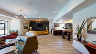 This single-floor space is designed as a sophisticated communal lounge and amenity area, combining open-plan living zones with a fully equipped kitchen. The layout flows seamlessly from one area to the next, unified by light hardwood-style flooring that runs throughout most of the space.

The lounge area is anchored by a bold dark geometric accent wall, which serves as a dramatic backdrop for a wall-mounted television and a cozy seating arrangement featuring mustard-yellow and striped accent chairs. A wood-paneled tray ceiling adds warmth overhead, complemented by a stylish multi-bulb chandelier. Nearby, a separate sitting area includes a fireplace framed by neutral plaster walls, bold orange upholstered chairs, and large glass doors that offer views and direct access to an outdoor pool area.

The kitchen is well-appointed with dark espresso cabinetry, light quartz countertops, stainless steel appliances including a full-size refrigerator, and a commercial beverage station. A striking abstract watercolor mural decorates one wall, adding an artistic focal point. A long bar-height island with bar stools provides additional seating and workspace. Recessed lighting and tray ceiling details with warm wood accents complete the refined, contemporary aesthetic throughout.