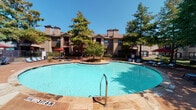 Located at 4604 Spruce Dr in Arlington, Texas, this single-floor residence offers a well-appointed interior complemented by impressive community amenities. The kitchen is a standout feature, showcasing dark granite countertops, a tile backsplash, light-toned cabinetry, stainless steel appliances, and wood-look flooring that adds warmth and durability. A breakfast bar provides a convenient transition between the kitchen and the adjoining living area, which is finished with plush carpet and crown molding throughout. The living space is brightened by windows fitted with blinds, and a ceiling fan with a light fixture ensures year-round comfort. The bedroom features mirrored closet doors that reflect light and enhance the sense of space, along with the same quality carpeting found throughout the home. Neutral, warm wall tones create a clean and inviting atmosphere across all rooms. Beyond the unit, residents have access to a generously sized outdoor swimming pool surrounded by a stone-tiled deck, lounge chairs, and shaded umbrella seating. Mature trees and well-maintained landscaping frame the pool area, with the multi-story residential buildings providing an attractive backdrop under the clear Texas sky.