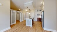 Located at 5350 Pinehurst Park Dr in Charlotte's desirable 28211 zip code, this single-floor residence offers a thoughtfully designed open-concept layout finished with warm, light-toned hardwood-style flooring that flows seamlessly throughout the space. The kitchen serves as the heart of the home, anchored by a generously sized center island with a white quartz countertop, an integrated sink, and elegant pendant lighting above. Cabinetry is finished in a striking two-tone combination — crisp white upper cabinets with glass-front accents reaching toward the ceiling, paired with rich warm-wood lower cabinets. Stainless steel appliances, including a full-size refrigerator, microwave, and range, complete the well-appointed culinary space. A tray ceiling detail above the kitchen adds architectural interest and a sense of volume. Sliding frosted-glass barn doors provide a stylish and functional transition to adjacent rooms, contributing a modern yet classic design element. The bedroom features neutral tones, recessed lighting, and access to a dedicated walk-in closet. The full bathroom is finished with large-format grey marble-look tile on both the floor and tub surround, complemented by a white vanity with quartz countertops and brushed nickel hardware, lending a clean and refined aesthetic throughout.