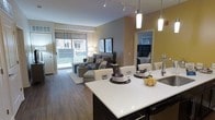 This single-floor residence showcases a thoughtfully designed open-concept layout that seamlessly connects the kitchen, dining, and living areas into one cohesive and inviting space. The kitchen is anchored by a striking dark-cabineted island with a crisp white countertop, complemented by elegant pendant lighting that adds a warm, modern ambiance. A dining area sits adjacent to the island, furnished with white upholstered chairs and a well-appointed table setting. The living room flows naturally from the dining space, featuring rich hardwood-style flooring, a comfortable sectional sofa, and layered décor including framed wall art and lush greenery. Track lighting runs along the ceiling, providing flexible illumination throughout the open area. A large sliding glass door floods the living space with natural light and opens to an outdoor balcony, offering views of the surrounding exterior. The bedroom, visible through a mirrored doorway, features a boldly upholstered headboard and continues the residence's contemporary aesthetic. Neutral wall tones and warm accent lighting throughout create a polished, welcoming atmosphere well-suited for both relaxing and entertaining.
