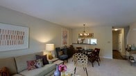 This two-floor residence offers a thoughtfully designed interior defined by neutral tones and a welcoming ambiance. The main living area is an open-concept space where the living room flows naturally into the dining area, creating a cohesive and functional layout. The living room is furnished with a comfortable sofa accented by decorative pillows, complemented by a classic table lamp that adds a warm, inviting glow to the space. Soft, light-toned carpeting extends throughout, enhancing the overall sense of warmth.

The dining area serves as a natural focal point, anchored by a round dining table surrounded by dark wood chairs and a woven accent chair. Above, a multi-arm chandelier provides an elegant lighting statement. Decorative wall art and signage add character to the dining space. Adjacent to the dining area, a kitchen pass-through counter with dark countertops connects seamlessly to the kitchen, allowing natural light from the kitchen window to filter into the main living space. A textured ceiling adds subtle architectural detail throughout. Together, these elements create a balanced, comfortable living environment spread across two floors.