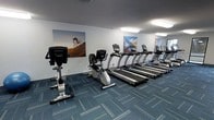 This single-floor fitness center is designed to support a focused cardio workout experience. The room is outfitted with a lineup of True-brand treadmills arranged in a row along one wall, complemented by both an upright and a recumbent stationary bike. The equipment is modern and well-maintained, catering to a range of fitness levels and preferences.

The space features a patterned blue-grey carpet that adds a clean, professional feel underfoot while providing practical durability. Overhead, flush-mounted panel lighting delivers consistent, even illumination throughout the room, creating a bright and energizing atmosphere without harsh shadows.

The walls are kept simple in a neutral white-grey tone, accented by large-format motivational photographs depicting high-intensity outdoor activities such as mountain biking and alpine landscapes. These bold visuals contribute to an energizing ambiance and serve as visual inspiration during workouts.

The layout is efficiently organized, allowing clear pathways between machines. Overall, the fitness center presents a tidy, functional environment well-suited for daily cardio exercise.
