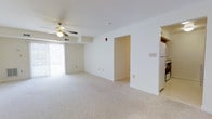 Located at 100 Golden Lane in Dover, Delaware, this single-floor residence offers a clean, functional layout designed for comfortable everyday living. The main living area is generously proportioned, featuring plush wall-to-wall carpeting that adds warmth underfoot. A ceiling fan with a built-in light fixture is centrally mounted, promoting air circulation and providing ambient lighting throughout the space. Large sliding glass doors along one wall flood the room with natural light and offer direct access to an outdoor area, creating a bright and airy atmosphere. The crisp white walls and low-profile ceiling design give the space a simple, well-maintained aesthetic. A clearly defined doorway leads to additional interior rooms, ensuring a logical and accessible flow throughout the home. Adjacent to the living area, the kitchen is partially visible and includes standard appliances, including a full-size refrigerator, positioned for convenient access. Structural elements such as a ceiling soffit add subtle architectural definition to the open plan. The overall ambiance is neutral and move-in ready, offering a versatile backdrop well-suited to a variety of interior styles and furnishing preferences.