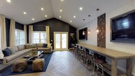 This single-floor space combines bold design choices with an open, inviting layout ideal for gathering and entertainment. The centerpiece of the room is a dramatic vaulted ceiling that peaks at impressive heights, fitted with recessed lighting that illuminates the space evenly throughout. Deep charcoal accent walls create a striking contrast against the lighter tones of the vaulted ceiling and trim, lending the room a sophisticated, modern edge.

A generously sized bar counter runs along one side of the room, accompanied by a row of industrial-style bar stools and accented by pendant lighting in warm metallic finishes. A decorative textured wallcovering with a botanical motif provides an eye-catching backdrop behind the bar area. A wall-mounted flat-screen television completes this entertainment zone.

On the opposite side, a comfortable lounge area features a plush sectional sofa and coffee table arranged atop a large area rug, creating an intimate seating space. French doors with cream-painted frames open to the outdoors, flooding the room with natural light. The flooring throughout is a light-toned herringbone wood, adding texture and elegance to the overall design. Together, these elements create a well-appointed, versatile space suited for both casual relaxation and social events.
