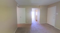 This single-floor residence features a straightforward, functional layout with a calm, neutral aesthetic throughout. The main living area is finished with wall-to-wall carpet in a muted tone, contributing to a quiet and comfortable atmosphere. Walls are painted in a warm beige, creating a consistent and understated color palette across the space.

A set of bifold closet doors provides built-in storage directly within the living area, offering convenient organization without occupying additional floor space. A ceiling-mounted air vent is visible, indicating a central climate control system integrated into the unit.

An open doorway connects the main living area to an adjoining room, which benefits from notable natural light streaming through a window. This secondary space appears to include cabinetry along the walls, suggesting it functions as a kitchen or utility area. The bright illumination from that room contrasts with the more subdued lighting of the main living space, drawing the eye through the open passage.

The textured ceiling adds a subtle visual detail overhead, while clean white baseboards frame the perimeter of each room. Overall, the space presents a simple, move-in-ready environment with practical features and a neutral design foundation.