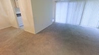 This single-floor space presents a straightforward and functional layout designed for comfortable everyday living. The main living area features wall-to-wall carpeting in a neutral beige tone, providing a soft underfoot feel that adds warmth to the interior. Smooth, light-colored walls keep the space feeling open and bright, while a structural column adds a subtle architectural element to the room's layout. A large window dressed with vertical blinds serves as the focal point of the living area, allowing an abundance of natural light to filter in and illuminate the space throughout the day. The blinds offer practical light control, enabling occupants to adjust brightness and privacy as needed. The overall ambiance is clean, neutral, and move-in ready, with a simple palette that provides a versatile backdrop for a variety of furnishing styles. The open feel of the carpeted area suggests generous usable floor space, making it well-suited for a comfortable seating arrangement. The understated design elements throughout contribute to a calm and livable atmosphere.