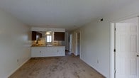 Located at 2100 Country Club Rd in Jacksonville, NC, this single-floor residence offers a well-organized and functional layout designed for comfortable everyday living. The open-concept design seamlessly connects the living area to the kitchen, creating a spacious and cohesive feel throughout the main living space. Soft carpeting covers the living area floor, adding warmth and comfort underfoot.

The kitchen is a standout feature, equipped with rich dark wood cabinetry that provides ample storage, a breakfast bar peninsula ideal for casual dining or meal prep, and a pendant light fixture overhead that adds a touch of character. A window above the sink allows natural light to filter into the kitchen, keeping the space bright and welcoming. The kitchen also includes essential appliances such as a refrigerator and range with an overhead microwave.

A textured ceiling adds subtle visual interest throughout the space. A hallway extends from the main living area, leading to additional rooms and revealing well-designed built-in closet shelving, offering generous and organized storage options. HVAC vents are positioned throughout to ensure consistent climate control. Overall, the space presents a clean, practical, and comfortable environment well-suited for residents seeking an efficient single-level home.