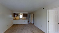Located at 2100 Country Club Rd in Jacksonville, NC, this single-floor residence offers a well-organized and functional layout designed for comfortable everyday living. The open-concept design seamlessly connects the living area to the kitchen, creating a spacious and cohesive feel throughout the main living space. Soft carpeting covers the living area floor, adding warmth and comfort underfoot.

The kitchen is a standout feature, equipped with rich dark wood cabinetry that provides ample storage, a breakfast bar peninsula ideal for casual dining or meal prep, and a pendant light fixture overhead that adds a touch of character. A window above the sink allows natural light to filter into the kitchen, keeping the space bright and welcoming. The kitchen also includes essential appliances such as a refrigerator and range with an overhead microwave.

A textured ceiling adds subtle visual interest throughout the space. A hallway extends from the main living area, leading to additional rooms and revealing well-designed built-in closet shelving, offering generous and organized storage options. HVAC vents are positioned throughout to ensure consistent climate control. Overall, the space presents a clean, practical, and comfortable environment well-suited for residents seeking an efficient single-level home.