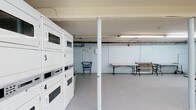 This single-floor laundry facility is designed with practicality in mind. The room features a bank of stacked washing machines and dryers along one wall, each unit clearly numbered for easy identification. The machines are finished in white, contributing to the clean, utilitarian aesthetic of the space. Overhead fluorescent lighting ensures the area is well-lit throughout.

A central structural column divides the open floor plan, while the light-painted concrete block walls reinforce the no-frills, functional character of the space. A folding table is positioned toward the center-right of the room, providing a convenient surface for sorting and folding laundry. A wooden bench with a slatted seat offers a place to sit while waiting. A wheeled laundry cart is also available near the machines for added convenience.

The concrete floor and exposed utility elements, including visible piping and a fire alarm, reflect the straightforward, utilitarian design of the facility. The open layout provides ample room to move around comfortably, making this a well-equipped shared laundry space.