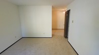This single-floor residential space offers a clean, straightforward layout designed for comfortable everyday living. The interior is finished with wall-to-wall carpeting in a warm beige tone throughout, providing a soft and cohesive feel underfoot across the rooms. Neutral white walls keep the space feeling bright and open, offering a versatile backdrop that adapts well to a variety of furnishing styles and décor choices.

The rooms are connected through a practical corridor layout, with a doorway visible between the main living area and an adjoining room, allowing for a natural flow through the space. Dark baseboards provide a subtle contrast against the light-toned walls and carpet, adding a grounding visual element to the interior.

A ceiling fan with a light fixture is installed in the adjoining room, contributing to both air circulation and ambient lighting. A light switch is conveniently positioned near the entryway between rooms. The overall ambiance is understated and functional, making this a practical space well-suited for a range of uses. The clean lines, neutral palette, and consistent flooring throughout contribute to a unified and easy-to-maintain interior environment.