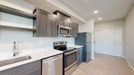 This well-designed single-floor unit offers a thoughtful layout that maximizes every square foot of its compact footprint. The open-concept kitchen and living area form the heart of the space, finished with light wood-style flooring and neutral gray walls that create a clean, contemporary aesthetic throughout.

The kitchen is a standout feature, equipped with a full suite of stainless steel appliances including a range, over-the-range microwave, dishwasher, and refrigerator. Rich, dark-toned shaker-style cabinetry with sleek hardware is paired with crisp white countertops, offering both style and functionality. Recessed lighting illuminates the kitchen workspace effectively.

The living area is bright and open, benefiting from natural light through windows and a glass-paneled exterior door that leads to a private balcony, providing outdoor views and additional ventilation. A wall-mounted HVAC unit ensures year-round climate control.

A separate bedroom area includes a sliding-door closet with built-in shelving, providing practical storage. The bathroom is neatly appointed with a dark-toned vanity with white countertop, a mirrored medicine cabinet, and a full tub-shower combination with clean white surround. Consistent light wood-style flooring flows throughout the entire unit, tying the spaces together cohesively.