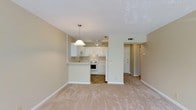 This single-floor space offers an open-concept design that seamlessly connects the living area and kitchen, creating a cohesive and functional layout. The living area features light-toned carpeting that lends a warm, understated feel, while a ceiling fan provides practical comfort overhead.

The kitchen is defined by crisp white cabinetry that extends to the upper walls, complemented by a breakfast bar that serves as a natural dividing element between the two spaces. A pendant light hangs above the bar, adding a focused decorative touch. White appliances, including a range and refrigerator, are integrated neatly into the cabinetry. The kitchen floor transitions to tile, offering a durable and easy-to-clean surface in the cooking area.

A hallway visible from the main living space leads to additional areas of the unit, with a entry door featuring a dark finish providing a subtle contrast to the otherwise light and neutral palette. Walls are finished in a soft beige tone throughout, contributing to the space's calm and airy ambiance. The textured ceiling and consistent trim work complete the clean, uniform aesthetic of this well-organized layout.