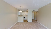 This single-floor space offers an open-concept design that seamlessly connects the living area and kitchen, creating a cohesive and functional layout. The living area features light-toned carpeting that lends a warm, understated feel, while a ceiling fan provides practical comfort overhead.

The kitchen is defined by crisp white cabinetry that extends to the upper walls, complemented by a breakfast bar that serves as a natural dividing element between the two spaces. A pendant light hangs above the bar, adding a focused decorative touch. White appliances, including a range and refrigerator, are integrated neatly into the cabinetry. The kitchen floor transitions to tile, offering a durable and easy-to-clean surface in the cooking area.

A hallway visible from the main living space leads to additional areas of the unit, with a entry door featuring a dark finish providing a subtle contrast to the otherwise light and neutral palette. Walls are finished in a soft beige tone throughout, contributing to the space's calm and airy ambiance. The textured ceiling and consistent trim work complete the clean, uniform aesthetic of this well-organized layout.