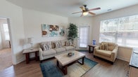 Located at Cagan Crossings in Clermont, Florida, this single-floor unit offers a thoughtfully designed layout with a warm, neutral palette throughout. The living area features wood-style flooring, a ceiling fan with light fixture, and multiple windows with slatted blinds that invite an abundance of natural light. A comfortable seating arrangement anchors the space around a stone-top coffee table, complemented by a colorful area rug and decorative wall art.

Adjacent to the living room is a dedicated dining nook with a round table and seating, paired with a wall mirror that enhances the sense of space. The kitchen is outfitted with dark espresso cabinetry, granite-style countertops, and white appliances, with a conveniently located in-unit laundry closet nearby.

The bedroom is carpeted and bright, featuring ceiling fan lighting, multiple windows, and direct access to an exterior door, lending an airy, private feel. A versatile secondary room serves as a home office or flex space, with wood-style flooring, a work desk, and ample natural light.

Both bathrooms are finished with dark cabinetry, granite countertops, tile flooring, and large mirrors. The overall ambiance is clean, bright, and functional — well-suited for comfortable everyday living in the Clermont community.