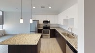 This single-floor unit presents a contemporary design with a cohesive neutral palette throughout. The open-concept kitchen and living area serves as the centerpiece, showcasing a thoughtfully designed kitchen equipped with full stainless steel appliances, including a refrigerator, electric range, and built-in microwave. Dark espresso lower cabinetry is paired with crisp white upper cabinets, while light granite countertops extend across the workspace and onto a central island, which features pendant lighting above. Light wood-look flooring runs throughout the main living area, enhancing the clean, modern aesthetic.

The living area adjoins the kitchen seamlessly and includes louvered utility closet doors, roller shades, and access to natural light through large windows. The bedrooms feature plush carpeting, floor-to-ceiling windows fitted with blackout roller shades, and baseboard heating units. Notable urban views are visible through the bedroom windows, suggesting an elevated position within a larger building.

The bathrooms maintain a consistent modern style, each featuring a full soaking tub with shower, a dark-cabinetry vanity with a light countertop, tile-look flooring, and clean white wall surrounds, creating a functional and streamlined finish.