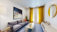 This single-floor residence offers a cohesive and stylish living experience with a carefully curated interior design throughout. The living area makes a strong first impression, featuring white walls that serve as a canvas for bold, colorful abstract artwork. Vibrant golden-yellow curtains frame a set of glass doors, flooding the room with natural light, while a patterned blue area rug and gold-accented coffee table add layers of visual interest. The kitchen is well-equipped and modern, showcasing a two-tone cabinetry design that pairs crisp white uppers with warm natural wood lower cabinets. Stainless steel appliances, including a French door refrigerator, range, and over-the-range microwave, provide a sleek, functional workspace. A peninsula with a white quartz countertop and pendant lighting above serves as both a prep area and a casual dining spot with stylish bar seating. The bedrooms offer a calm and restful atmosphere, each finished with plush carpeting, recessed lighting, and clean white walls. One bedroom features access to a walk-in closet, while both rooms provide ample space and a warm, welcoming feel. The bathroom is clean and contemporary, with a tiled tub-shower combination, a wood-toned vanity with a white countertop, and light tile flooring that ties the space together cohesively.