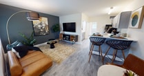 This single-floor unit offers a thoughtfully designed open-concept layout that seamlessly connects the living area and kitchen. The living space features light wood-style flooring throughout, a bold dark accent wall that serves as an eye-catching focal point, and ample room for comfortable furnishings. A wall-mounted flat-screen television anchors the living area, complemented by warm, modern décor elements.

The kitchen is efficiently arranged with gray shaker-style cabinetry, white countertops, and a tiled backsplash. A peninsula with bar seating for two provides a casual dining option and reinforces the open flow between the kitchen and living space. Stainless steel appliances, including a refrigerator and microwave, complete the kitchen's functional design.

The bedroom is a calm, neutral-toned retreat featuring the same light wood-style flooring. A wide horizontal window frames lush greenery and palm foliage outside, allowing natural light to fill the room. An open closet provides organized storage space, and an adjoining bathroom is conveniently accessible directly from the bedroom. The overall ambiance of the unit is clean, contemporary, and inviting.
