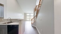 This well-appointed two-floor unit spans 830 square feet, with 630 square feet on the main level and 200 square feet on the upper level, connected by a wood-railed staircase. The main level boasts soaring vaulted ceilings and large multi-pane windows that flood the living area with natural light and provide treetop views of the surrounding greenery. Durable grey wood-look luxury vinyl plank flooring runs throughout the main living spaces, complementing the neutral warm-grey wall tones. The kitchen is equipped with grey shaker-style cabinetry, light stone countertops, and black appliances including a refrigerator, range, and over-the-range microwave. A full bathroom on the main level features a tub-shower combination with white tile surround, a grey vanity with countertop, and matching LVP flooring. The upper level, accessible via the staircase, offers a carpeted bedroom area with a double-door closet and recessed lighting. Additional rooms feature LVP flooring, ample wall outlets, and windows overlooking outdoor views. The unit's clean, modern aesthetic and thoughtful layout make it a functional and comfortable living space.