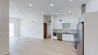 This contemporary single-floor space showcases a clean, open-concept design that seamlessly blends the living and kitchen areas into one cohesive environment. The kitchen is thoughtfully appointed with crisp white cabinetry — including both upper and lower units — complemented by dark countertops that provide a striking contrast. Stainless steel appliances, including a dishwasher, oven, and refrigerator, lend a polished, modern aesthetic. A frosted glass cabinet panel adds a subtle decorative touch among the upper cabinetry, while a stainless steel sink and tiled backsplash round out the functional workspace. Light-toned hardwood flooring extends throughout the open living area, enhancing the sense of spaciousness and warmth. Recessed lighting is thoughtfully distributed across the ceiling, ensuring even illumination throughout the space. The high ceilings amplify the airy, expansive feel of the interior. Neutral white walls provide a versatile backdrop, while strategically placed HVAC vents ensure year-round comfort. A hallway visible in the background hints at additional areas beyond the main open floor plan, including what appears to be a closet or storage space. Overall, the space exudes a minimalist, move-in-ready appeal with high-quality finishes.