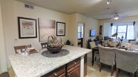 This single-floor space showcases an open-concept design that seamlessly connects the kitchen, dining, and living areas. The kitchen is anchored by a large granite-topped island with rich, dark wood cabinetry beneath, offering both generous counter space and additional storage. The light-toned granite surface features subtle veining, adding an elegant touch to the space.

The open layout allows for a natural flow into the dining area, which comfortably accommodates a full dining set with upholstered seating. Warm, neutral-toned walls throughout create a cohesive and inviting ambiance, complemented by decorative framed artwork that adds visual interest.

The living area is visible beyond the dining space, furnished with comfortable seating and anchored by a wall-mounted flat-screen television. A window in the background allows natural light to filter in, brightening the interior. Crown molding along the ceiling adds a refined architectural detail throughout the space. The overall design balances functionality with aesthetic appeal, combining warm wood tones, soft neutrals, and quality finishes to create a welcoming and practical living environment.