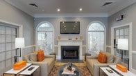 This single-floor clubhouse is designed to provide a welcoming and sophisticated community gathering space. The centerpiece of the main lounge is a classic white-mantled fireplace flanked by two dramatic arched windows with decorative fanlights, filling the room with natural light. A large flat-screen television is mounted above the mantel, making the space ideal for both relaxed socializing and group viewings. Neutral gray walls, crown molding, and white wainscoting throughout lend a refined, traditional character to the interior, while warm hardwood flooring transitions seamlessly between areas.

The seating area features plush beige upholstered chairs accented with vibrant orange throw pillows, arranged around a glass-top coffee table atop a geometric patterned area rug, creating an inviting and cohesive aesthetic. Bold abstract artwork in matching orange and white tones further ties the color palette together.

A casual dining nook with a round wood table and modern gray chairs provides a more intimate setting, complemented by additional abstract wall art. Adjacent to this area, a refreshment station near the exterior door offers snacks and beverages, conveniently positioned for residents on the go. A windowed pass-through connects the lounge to a private office area, ensuring staff accessibility while maintaining an open, airy flow throughout the clubhouse.