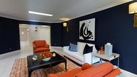 Located at 485 S Parkview Ave in Columbus, Ohio, this single-floor property makes a strong design statement through its bold and carefully curated interior aesthetic. The living area showcases a dramatic color palette centered on deep navy blue walls that create a rich, sophisticated backdrop for the space. Crown molding runs along the ceiling, adding a refined architectural detail that elegantly frames the room. The interior is brought to life with vibrant coral and white upholstered seating, including a plush sofa adorned with navy and blue accent pillows, and complementary armchairs that reinforce the cohesive color story. A large-scale black and white abstract artwork serves as a commanding focal point on the navy feature wall, adding an artistic and contemporary flair. The space is anchored by a geometric orange and white area rug beneath a sleek black lacquered coffee table, which ties the bold tones together harmoniously. Warm wall sconces with gold fixtures provide ambient lighting, enhancing the inviting yet refined atmosphere. The overall ambiance of this property is one of curated elegance — bold, intentional, and visually dynamic, making it a distinctive and memorable space.