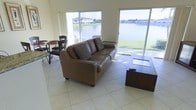This two-floor space offers a welcoming and open layout designed to maximize comfort and natural light. The main living area is anchored by large sliding glass doors that frame expansive waterfront views, creating a seamless connection between the interior and the lush, green outdoor surroundings. Light-toned tile flooring runs throughout, contributing to the bright and airy atmosphere of the space.

The living area is generously proportioned, accommodating a full leather sofa ensemble and a dedicated dining area with table and chairs, making it well-suited for both relaxing and entertaining. The sliding glass doors provide direct access to the exterior, where a serene waterfront setting with manicured landscaping can be enjoyed.

Natural light pours in from multiple directions through large windows and glass doors, keeping the interior feeling open and connected to the outdoors throughout the day. The neutral color palette of the walls and floors complements the natural scenery visible from within, reinforcing a calm and refreshing ambiance.

Spread across two floors, this space offers versatile living arrangements with ample room to accommodate a variety of needs. The overall design prioritizes views, light, and an open, functional layout that makes the waterfront setting a central feature of the everyday living experience.