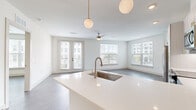 This well-appointed 1-bedroom, 1-bathroom residence offers a thoughtfully designed open-concept layout that maximizes both space and functionality. Light-toned luxury vinyl plank flooring flows seamlessly throughout, complementing the crisp white walls and creating a clean, cohesive aesthetic.

The combined kitchen and living area serves as the heart of the home. The modern kitchen features two-tone cabinetry — white upper cabinets paired with soft blue-grey lower cabinets — accented with gold hardware. A peninsula with a white quartz countertop, undermount sink, and stylish globe pendant lights anchors the space. Stainless steel appliances, including a refrigerator, range, microwave, and dishwasher, complete the kitchen. An in-unit washer is conveniently tucked nearby.

The living area is bathed in natural light through multiple large windows and French doors that open to an outdoor view, enhanced by a ceiling fan overhead. The bedroom offers a comfortable footprint with its own window and ceiling fan, while a dedicated closet provides practical storage.

The bathroom showcases the same blue-grey cabinetry with gold accents found in the kitchen, maintaining a unified design language. A private hallway entry and recessed lighting throughout add polished finishing touches to this bright, modern residence.