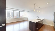 Located in the heart of downtown Buffalo at 10 Lafayette Square, this thoughtfully designed one-bedroom, two-bathroom residence offers a refined urban living experience. The open-concept kitchen and living area serves as the centerpiece of the unit, where rich hardwood flooring flows throughout and floor-to-ceiling windows frame sweeping views of the Buffalo skyline, flooding the space with natural light. The kitchen is equipped with stainless steel appliances, including a refrigerator, electric range, and built-in microwave, complemented by rich dark cabinetry, white countertops, and a large central island with pendant lighting overhead — ideal for both cooking and casual dining. The spacious primary bedroom continues the hardwood flooring and panoramic city views, featuring a ceiling fan and a dedicated closet. It connects to a well-appointed en-suite bathroom with a glass-enclosed walk-in shower finished in classic white subway tile. A second bathroom provides additional convenience for guests. Throughout the unit, a consistent modern aesthetic is maintained with dark-toned doors and cabinetry contrasting against crisp white walls, creating a clean, contemporary ambiance that complements its vibrant downtown Buffalo location.