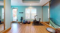 Located in La Quinta, California, this dedicated fitness studio is a thoughtfully designed workout space that combines functionality with stylish aesthetics. The room is finished with warm hardwood flooring throughout and painted in a calming aqua-blue palette that creates an energizing yet relaxed atmosphere. A tray ceiling with recessed lighting adds architectural depth and ensures the space is well-lit for exercise.

A striking floor-to-ceiling abstract mural in swirling shades of teal, white, and gold serves as a dramatic focal point on one wall, lending an artistic sensibility to the room. Large framed mirrors are strategically positioned along the walls to amplify the sense of space and support proper form during workouts. Natural light filters in through multiple windows fitted with blinds, offering a bright and inviting environment.

The studio is fully equipped with a wide array of fitness equipment, including an air resistance exercise bike, stability balls, a dumbbell rack, kettlebells, a medicine ball tower, resistance bars, yoga mats, and step platforms. Open shelving units provide organized storage for accessories and equipment. A wall-mounted television is also present, ideal for following workout classes or entertainment during training sessions. This space is a complete, private fitness retreat.